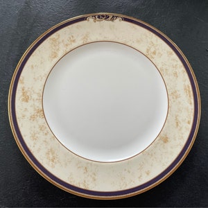 Puede incluir: Un plato de porcelana blanco con un borde color crema con motas doradas y una banda azul oscuro. El plato tiene un borde dorado y un diseño decorativo en la parte superior. Este elegante plato es adecuado para cenas formales.