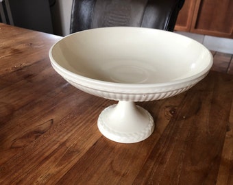 May include: A white ceramic pedestal bowl with a fluted rim and a braided design on the base.