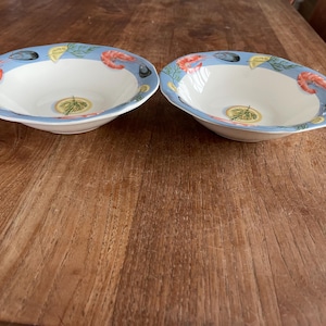 Könnte beinhalten: Zwei weiße Keramikschalen mit hellblauem Rand, die Illustrationen von Garnelen, Muscheln und Zitronenscheiben zeigen. Die Schalen haben einen gewellten Rand und ein zentrales Design einer Zitronenscheibe mit Kräutern. Auf einer Holzoberfläche.