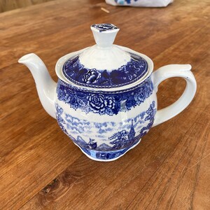 Könnte beinhalten: Weiße Keramik-Teekanne mit blauem Blumen- und Landschaftsdesign. Die Teekanne hat einen gebogenen Ausguss, einen abgerundeten Griff und einen Deckel mit einem floralen Knauf. Das blaue Design zeigt Rosen und eine Landschaftsszene.