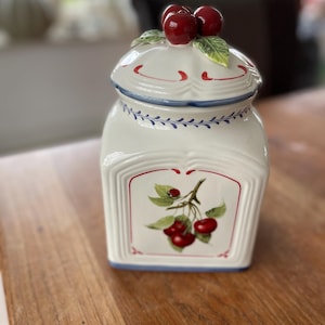 Puede incluir: Tarro de cerámica blanco con un diseño floral azul y blanco y una tapa con tres cerezas rojas en la parte superior. El tarro tiene una imagen de una rama de cerezo con cerezas rojas en ella.