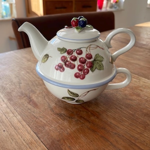Può includere: Un servizio da tè in ceramica bianca composto da una teiera e una tazza. La teiera presenta un coperchio con un grappolo di bacche e un manico. Il corpo della teiera e della tazza è decorato con un grappolo di ciliegie rosse e foglie verdi.