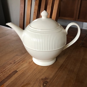 Può includere: Una teiera in ceramica bianca con un design a coste e un coperchio. La teiera è appoggiata su un tavolo di legno.