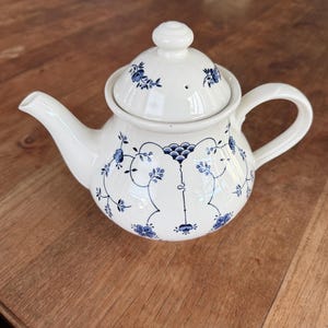 Peut inclure: Une théière en céramique de couleur crème ornée d'un délicat motif floral bleu composé de fines vignes et de petites fleurs. La théière comprend un couvercle assorti avec un bouton arrondi, un bec verseur courbé et une anse robuste.