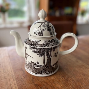 May include: A cream-coloured ceramic teapot featuring a dark brown transferware pattern of woodland scenes, including trees, foliage, and small animals. The teapot has a matching lid with a pointed finial, a curved handle, and a spout.