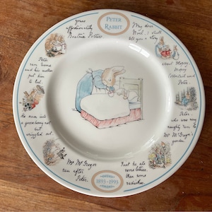 May include: A white ceramic plate featuring a blue and red illustration of Peter Rabbit in bed. The plate has a blue border with text around the edge, including "Peter Rabbit", "My dear Noel, I shall tell you a story", and "yours affectionately Beatrix Potter".