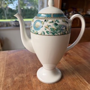 Op de afbeelding: Een witte porseleinen theepot met een voetstuk, een gebogen handvat en een tuit. Het bovenste gedeelte is versierd met een kleurrijke band van blauwgroene, groene en gele bloem- en vogelmotieven, geaccentueerd door een geometrische rand en een deksel met een gouden punt.
