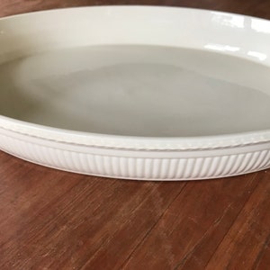 May include: White oval ceramic baking dish with a fluted rim. The dish is empty and sitting on a wooden surface.