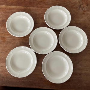 Puede incluir: Seis platos de cerámica blanquecina con bordes festoneados están dispuestos sobre una superficie de madera. Los platos tienen un diseño sencillo y elegante, adecuado para servir aperitivos o postres. Los platos son redondos con un borde ligeramente elevado.