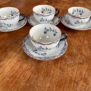Può includere: Quattro tazze e piattini bianchi con disegni floreali blu e manici blu sono disposti su una superficie di legno. I bordi delle tazze e dei piattini hanno un bordo blu e arancione.