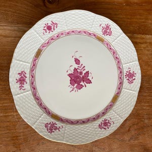 Può includere: Un piatto in porcellana bianca con un motivo floreale rosa e un bordo dorato. Il piatto ha un motivo intrecciato sulla superficie.