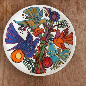 Op de afbeelding: Een rond wit keramieken bord met een kleurrijke illustratie van drie vogels tussen gestileerde bloemen en bladeren. Het ontwerp bevat tinten blauw, oranje, rood, paars en groen, met dikke zwarte lijnen die de vormen definiëren.