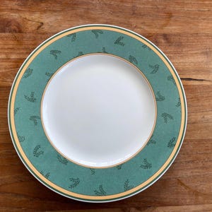Puede incluir: Un plato blanco redondo con un borde verde azulado con un patrón de hojas repetido. El borde está delineado con una fina banda dorada. El plato está sobre una superficie de madera.