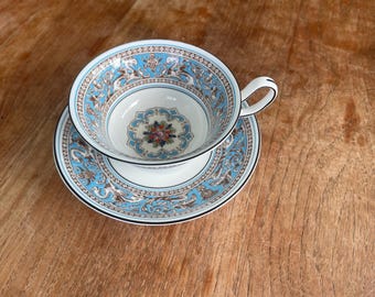 Può includere: Un set vintage di tazza e piattino da tè. La tazza e il piattino hanno una base bianca con un motivo floreale blu e marrone dettagliato. La tazza ha un manico curvo e il set è posto su una superficie di legno.