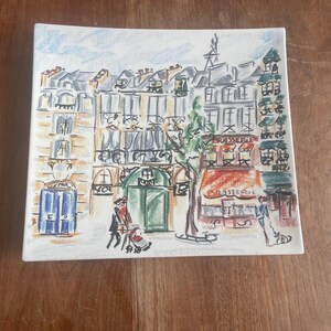 May include: A hand-painted ceramic plate depicting a charming Parisian street scene. The buildings are painted in shades of yellow, pink, and blue, with a green awning over a cafe labelled "Brasserie".