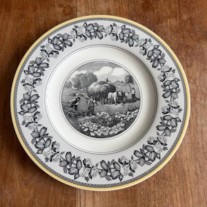 Puede incluir: Un plato de cerámica blanco con una escena pastoral en blanco y negro en el centro. El plato tiene un borde amarillo y un borde de diseños florales en blanco y negro. El plato está sobre una superficie de madera.