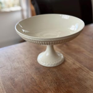 Pode incluir: Tigela de servir com pedestal creme, com uma tigela larga e rasa e uma base decorativa canelada. A tigela assenta num pedestal curto e arredondado com um design detalhado em relevo. A tigela é feita de cerâmica e está numa mesa de madeira.