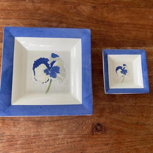 Op de afbeelding: Twee vierkante keramische schalen met een blauwe rand en een witte binnenkant. De schalen hebben een bloemenontwerp met blauwe en witte bloemblaadjes. De grotere schaal meet ongeveer 20 cm vierkant, de kleinere ongeveer 10 cm.