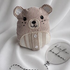May include: A stuffed bear toy made of brown and white striped fabric with embroidered black eyes, nose, and mouth. It features a cream-coloured belly panel with two white buttons. Beside it is a clear oval acrylic tag with the text blake + boden.