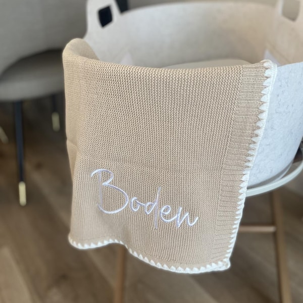 Könnte beinhalten: Eine beige gestrickte Babydecke mit weißer Einfassung und dem Namen "Boden" in weißer Schrift gestickt. Die Decke liegt über einer weißen Babywiege.