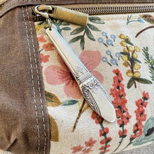 Może przedstawiać: Zbliżenie płóciennej torby z kwiatowym wzorem i srebrnym uchwytem zamka błyskawicznego w kształcie łyżki. Torba ma brązowy płócienny bok z białymi szwami.