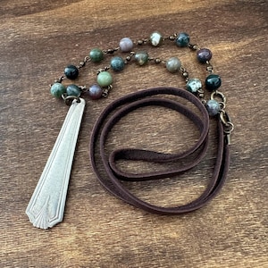May include: A necklace featuring a silver-toned metal spoon handle pendant attached to a dark brown leather cord. The cord is accented with a series of multi-colored round beads in shades of green, grey, and purple, connected by bronze-toned metal links.