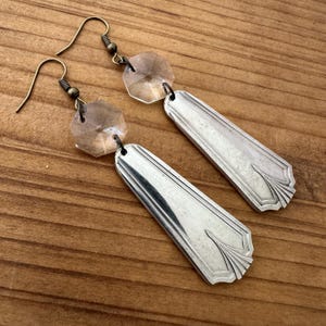 May include: A pair of dangle earrings featuring silver-toned metal spoon handle charms with embossed line detailing. Each earring is accented with a clear, faceted hexagonal glass bead attached to a dark metal hook for pierced ears.