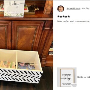 May include: A white and black chevron patterned book organizer with three compartments, filled with colorful children's books. A sign that says "baby" is on top of a wooden cabinet.