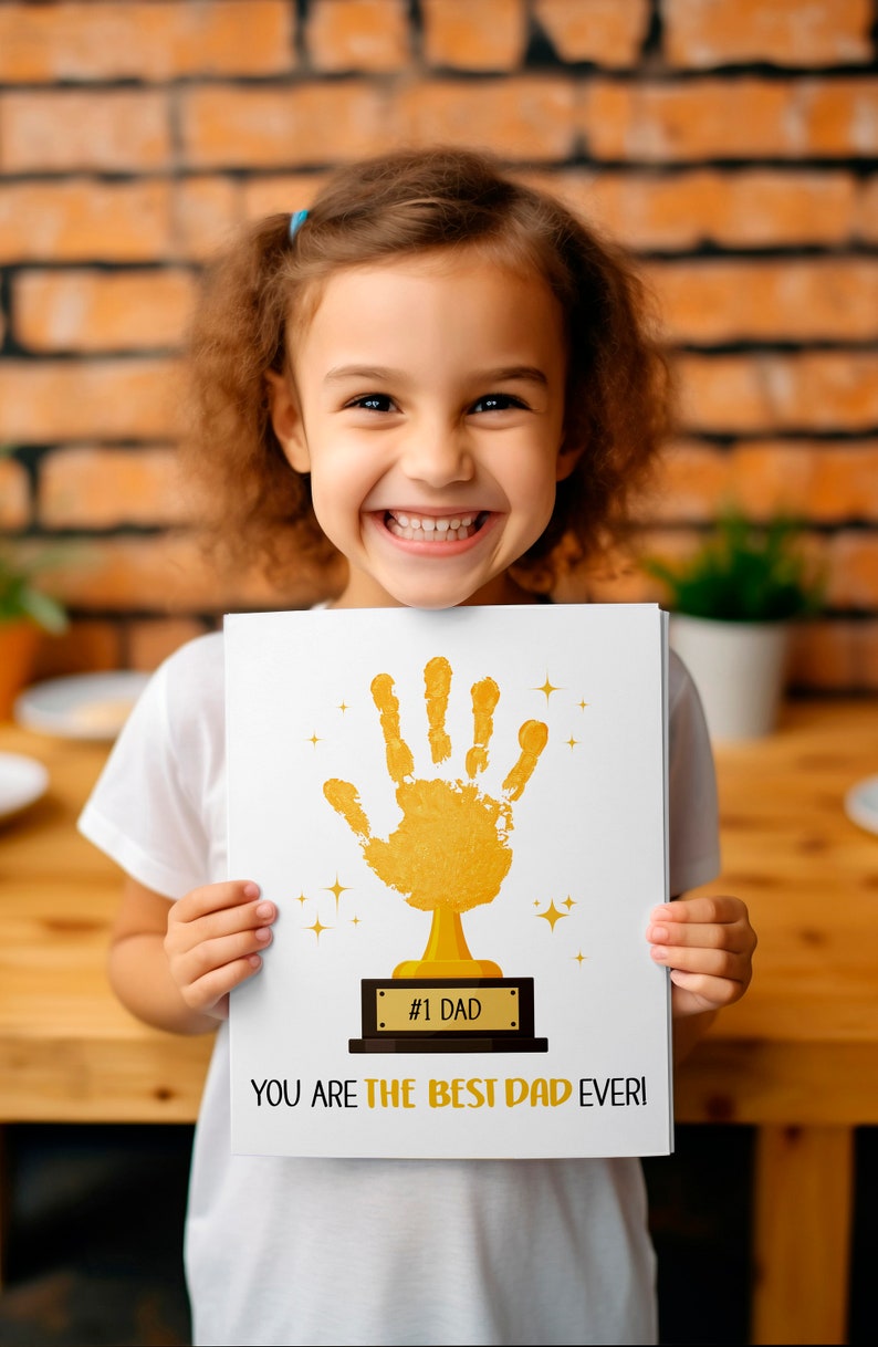 May include: A child holds up a white card with a gold handprint trophy on it. The card reads "#1 Dad You Are The Best Dad Ever!"