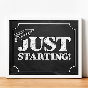 May include: Black chalkboard sign with a white frame. The sign has a white graduation cap drawn on it and the text "JUST STARTING!" in white chalk.