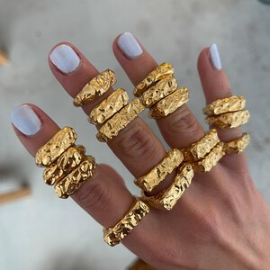 May include: A hand wearing multiple gold-toned chunky rings with a textured surface. The rings are stacked on each finger.