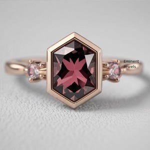 Könnte beinhalten: Ein Ring aus Roségold mit einem sechseckigen, tief roten Edelstein, der von zwei kleineren, rosafarbenen Edelsteinen umgeben ist. Der Ring liegt auf einem weißen Hintergrund. 'Eminent Jewels' ist in der oberen rechten Ecke des Bildes gedruckt.