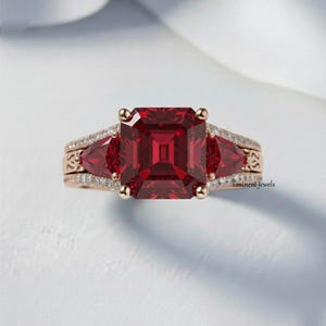 May include: A rose gold ring featuring a large, square-cut, deep red gemstone as the centerpiece, flanked by two smaller, triangular red gems. The band is adorned with small, sparkling white stones and intricate detailing.