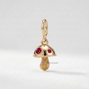 Può includere: Un ciondolo a forma di fungo in oro con finitura lucida e gambo testurizzato. Il cappello del fungo è decorato con due pietre preziose rotonde di colore rosso vivace incastonate nel metallo. In alto è presente una contromaglia e il testo Eminent Jewels.