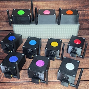 May include: A collection of black electronic devices with colored buttons, antennas, and power switches. The buttons are various colors, including green, pink, orange, blue, yellow, red, and white. The devices are arranged on a wooden surface.