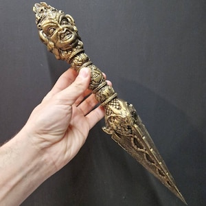 May include: A detailed, golden-colored dagger with an ornate handle featuring a sculpted face. The blade is pointed and has decorative patterns. The dagger is held against a dark background, showcasing its intricate design.
