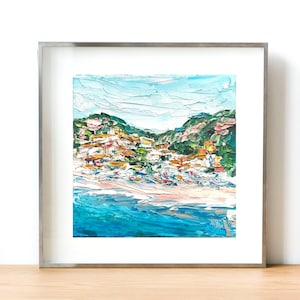 May include: A framed painting of a coastal town with colorful buildings on a hillside overlooking a blue ocean. The painting is done in a textured style with visible brushstrokes.
