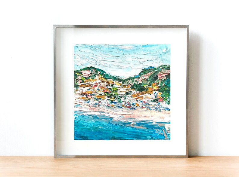 May include: A framed painting of a coastal town with colorful buildings on a hillside overlooking a blue ocean. The painting is done in a textured style with visible brushstrokes.