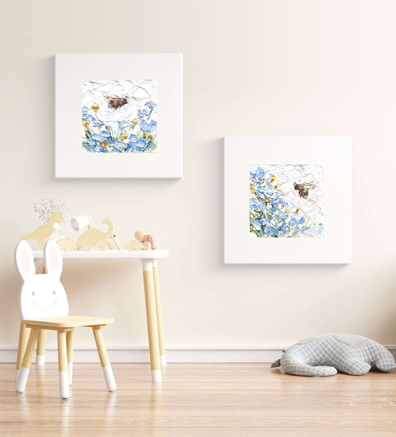 May include: Two framed paintings of blue flowers with bees flying around them. The paintings are hanging on a white wall in a room with a wooden table and chair.