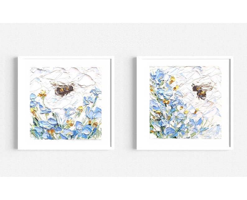 May include: Two framed paintings of a bee flying over blue flowers with yellow centers. The paintings are on a white wall.
