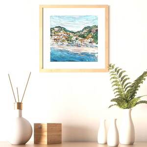 May include: A framed painting of a coastal town with colorful buildings on a hillside overlooking a blue ocean. The painting is in a light wood frame and is hanging on a white wall.
