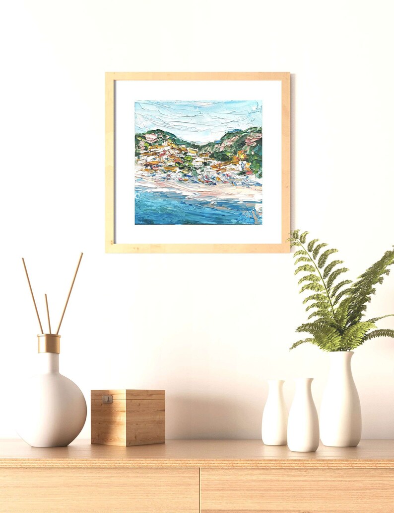 May include: A framed painting of a coastal town with colorful buildings on a hillside overlooking a blue ocean. The painting is in a light wood frame and is hanging on a white wall.