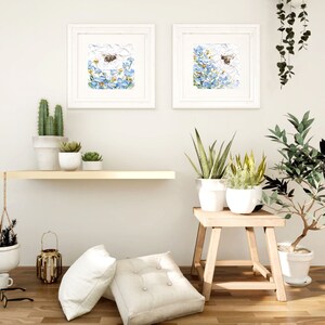May include: Two framed watercolor paintings of blue flowers with a bee flying above them. The paintings are hanging on a white wall above a wooden shelf with potted plants. A wooden stool with a potted plant sits in front of the shelf.
