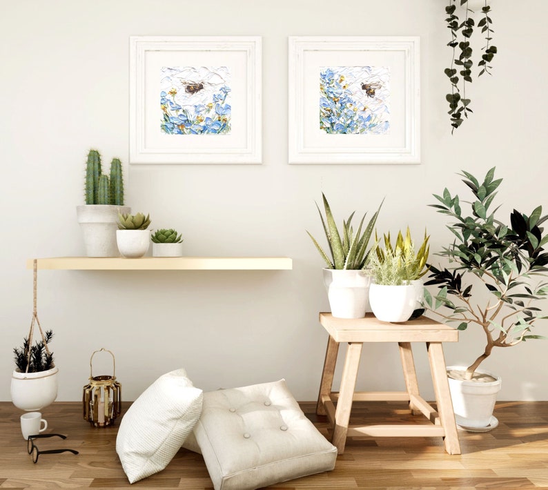 May include: Two framed watercolor paintings of blue flowers with a bee flying above them. The paintings are hanging on a white wall above a wooden shelf with potted plants. A wooden stool with a potted plant sits in front of the shelf.