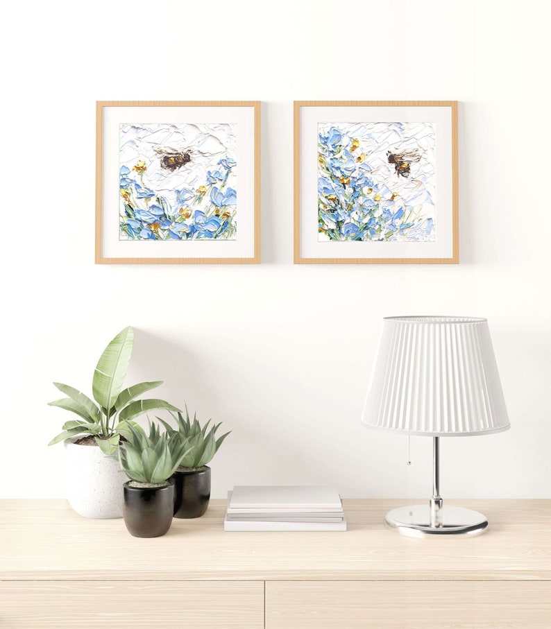 May include: Two framed paintings of blue flowers with a bee flying in front of them. The paintings are hanging on a white wall above a wooden table with a lamp and potted plants.