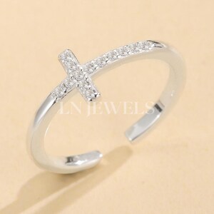 May include: A silver-toned ring with a cross design. The cross is adorned with small, clear gemstones. The ring band curves around the finger, with the text "LN JEWELS" visible on the ring.