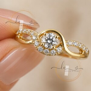 May include: A gold ring featuring a central round brilliant-cut clear stone surrounded by a partial halo of smaller pavé-set stones. The band has a crossover design with additional small clear stones embedded along the polished gold metal surface.