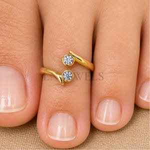 May include: A gold-colored toe ring with two clear, round gemstones. The ring wraps around the toe with a curved design. The text "JEWELS" is partially visible.