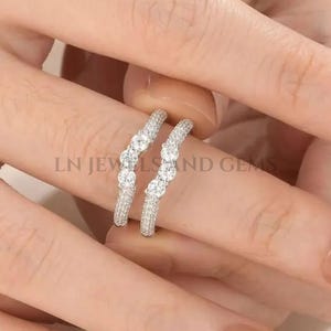 May include: Two silver-toned, curved rings with clear gemstones. The rings are set with multiple small stones and larger center stones. The rings are displayed on a light-colored surface. The text "LN JEWELS and GEMS" is visible.