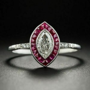 Puede incluir: Un anillo en forma de marquesa con un diamante central y un halo de piedras rojas. La banda del anillo es plateada y está adornada con pequeñas piedras preciosas transparentes. El anillo se presenta sobre un fondo oscuro, resaltando su brillo.